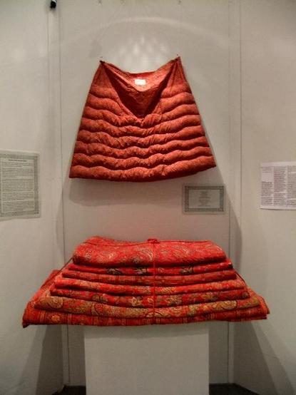 A red quilted skirt hanging up on a wall in an exhibition, with a pile of red paisley quilts beneath it.
