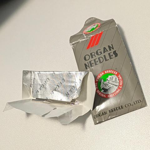 A silver gum-wrapper-style foil packet opened to expose sewing machine needles, next to an Organ Needles paper wrapper.