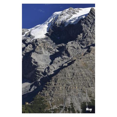 Portrait photo of a small section of the Ortler mountain, incl. fully glaciated summit region, gigantic bare rock faces, spurs, and a small mountain hut in the bottom left corner, near the treeline. Clear deep blue sky and shadows emphasizing the rock structures.