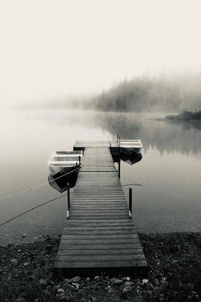Black and white photograph of two small boats on the water of a lake, hung on each side of a wooden dock. The pebble bank is visible at the bottom of the photo. The "t" shaped dock advances into the water to the middle of the photo, with metal posts and mooring lines installed on each side, as well as two ropes attached on each side, above the water and disappearing left and right. Boats and ropes reflect perfectly on the mirror surface of the lake. Behind, the opposite shore of the lake disappears almost completely in white fog. A hill covered with forest, a point of land and the white sky are reflected in the water.

Photographie noir et blanc de deux petits bateaux sur l'eau d'un lac, accrochés de chaque côté d'un quai en bois. La rive de galets est visible dans le bas de la photo. Le quai en forme de "t" s'avance dans l'eau jusqu'au milieu de la photo, avec des poteaux et amarres en métal installés de chaque côté, ainsi que deux cordes attachées de chaque côté, au-dessus de l'eau et disparaissant à gauche et à droite. Les bateaux et les cordes se reflètent parfaitement sur la surface miroir du lac. Derrière, la rive opposée du lac disparaît presque complètement dans la brouillard blanc. Une colline couverte de forêt, une pointe de terre et le ciel blanc se reflètent dans l'eau.
