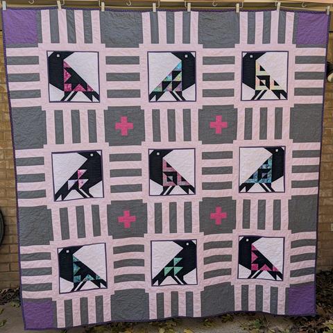 A square quilt featuring 9 crows with various quilt blocks as their wings.  The blocks are in bright colors.  The crow blocks are surrounded by lilac frames, with a grey background.  There are 4 hot pink crosses in the background blocks near the middle crow, and the 4 corners of the quilt are purple instead of grey.