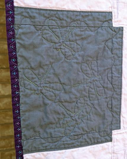 Dragonflies stitched in blue-green shiny thread on a grey background.  Next to them is a the quilt border of dark purple with multi-colored bird-foot-like decorative stitching.