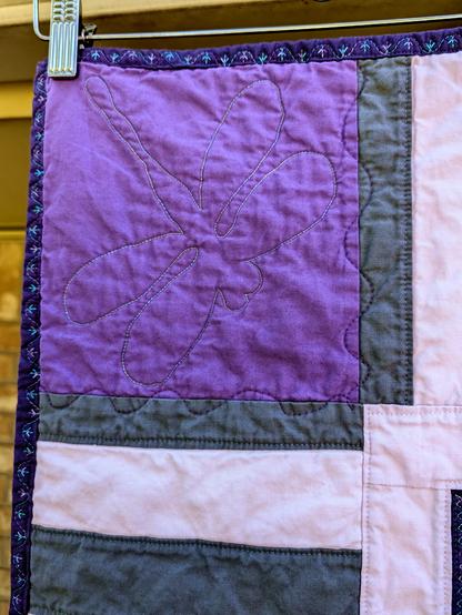 A big dragonfly stitched in blue-green shiny thread on a medium purple background.  Next to it is a the quilt border of dark purple with multi-colored bird-foot-like decorative stitching.