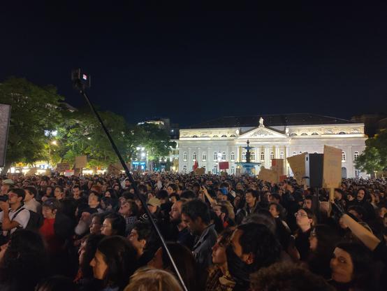 A square absolutely packed with hundreds of people, holding signs and looking to the left (likely towards the center of the square). The D Maria II theatre is lit up in the background, looking quite lovely.