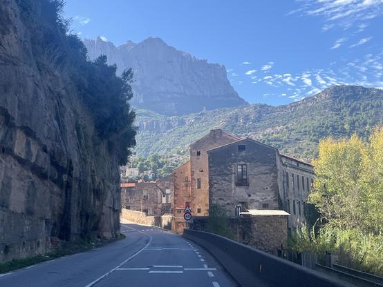 Orange-hued stone buildings are illuminated by the afternoon sun, as the Montserrat mountain looms behind under a blue sky