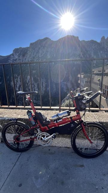 A red bicycle perches against a railing at the edge of the Abbey of Montserrat in Catalonia, with the afternoon sun set above the jagged mountaintop