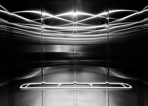 Black and white photograph of the interior of an elevator, showing walls covered with vertical rectangular aluminum panels, reflecting ceiling lights in a pattern of intertwined curved lines. A horizontal handrail is fixed lower on the wall.

Photographie noir et blanc de l'intérieur de la cage d'un ascenseur, montrant les murs couverts de panneaux d'aluminium rectangulaires verticaux, réfléchissant les lumières du plafond dans un pattern de lignes courbes entremêlées. Une main courante horizontale est fixée plus bas sur le mur.