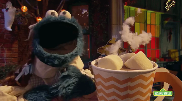 GIF of Cookie Monster in a dressed as Dorthy from The Wizard of Oz attempting to snatch a marshmallow out of a giant cup of hot cocoa that turns out to be Abby Cadabby in a Halloween costume, who scares Cookie Monster away