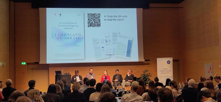 A panel of 5 people sit on a stage in a wooden lined room in front of a screen advertising a new report with a qr code to download it