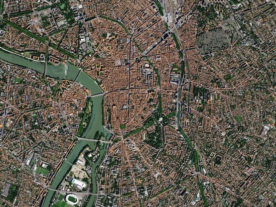 Dense historic city center with terracotta roofs and star-like radial streets converging on a central plaza, bounded by a winding river crossed by several bridges and surrounded by parks and dense residential neighborhoods.