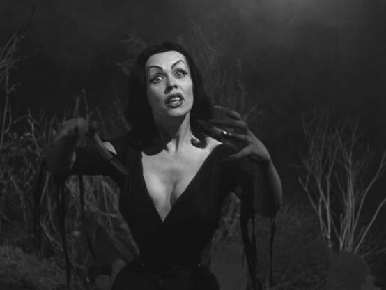 Maila Nurmi as Vampira in the 1957 film "Plan 9 from Outer Space". She's looking at us with her long black hair and slinky dress. Her arms are outstreched toward us. We're dooomed I tell ya!