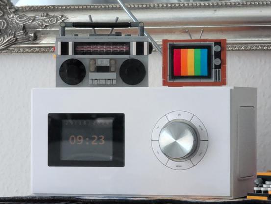 Die Aufnahme zeigt einen Mini-TV aus Klemmbausteinen, der sich durch sein Testbild in Regenbogenfarben deutlich vom weiss-schwarz-grau in seiner Umgebung abhebt...

Die Szene zeigt ein kantiges, quaderförmiges, weisses DAB/Internetradio mit nur schwach erkennbarer Uhrzeit + Datum auf dem Display; oben drauf steht links ein grau-schwarzer Mini-Ghetto-Blaster und rechts daneben besagter, bunter Mini-Fernseher aus Klemmbausteinen; dahinter ist noch anteilig der silberne Spiegelrahmen mit Ornamente erkennbar
