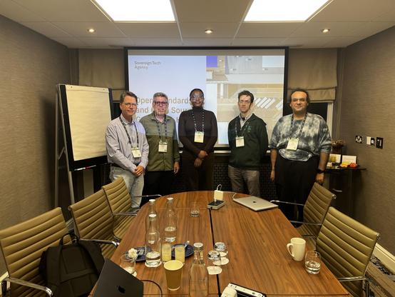 A group of five people posing for a photo in a conference room, standing behind a large wooden conference table with chairs and water glasses. A projection screen behind them displays text including "Sovereign Tech Agency".