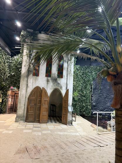 Eine Indoor-Szene mit einer verwitterten Kapelle mit Buntglasfenstern und Holztüren, umgeben von üppigem Grün. Der Boden ist sandig, mit Holzwegen, die zum Eingang führen. Zu den sichtbaren Elementen gehören Palmen und eine dunkle Wand, die zur tropischen Atmosphäre beitragen.