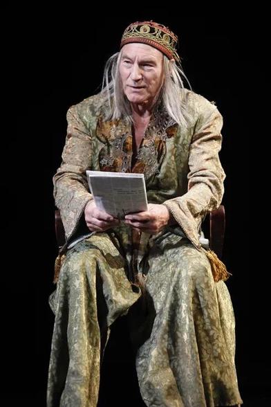 Stewart wearing a kind of long golden green gown while sitting in a chair in costume as King Lear